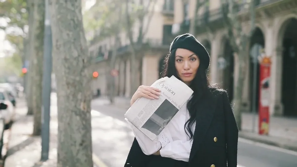 Elegant woman in a beret posing with a newspaper on a sunny street.